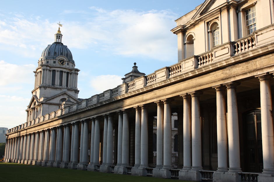 A historic white stone building with a prominent domed tower features a clock and decorative columns at its entrance. In front, a paved area includes several large cardboard moving boxes, some wrapped