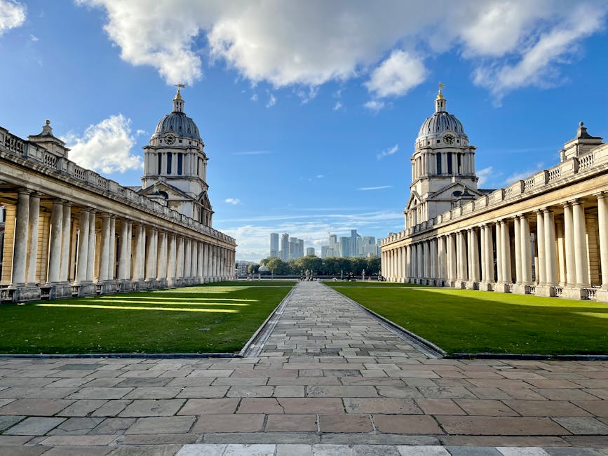 A wide shot of the historic Old Royal Naval College in Greenwich, London, showing two symmetrical, domed towers with classical architectural details made of stone, flanking a central pathway paved with rectangular stones. The pathway leads towards a distant view of modern skyscrapers against a partly cloudy blue sky. On either side of the pathway, there are well-maintained green lawns bordered by a colonnade with evenly spaced columns supporting a balustrade. The scene is illuminated by natural daylight, highlighting the detailed stonework of the buildings and the contrast between the historic architecture and the city skyline in the background. This image captures the grandeur of the historic structure, often associated with iconic London landmarks, and can be related to urban relocation or sightseeing visits to Greenwich.