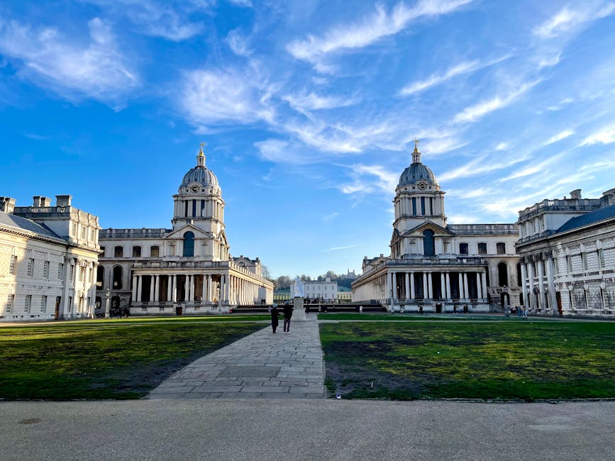 A wide shot of the historic Old Royal Naval College in Greenwich, London, showing two symmetrical, domed towers with classical architectural details made of stone, flanking a central pathway paved with rectangular stones. The pathway leads towards a distant view of modern skyscrapers against a partly cloudy blue sky. On either side of the pathway, there are well-maintained green lawns bordered by a colonnade with evenly spaced columns supporting a balustrade. The scene is illuminated by natural daylight, highlighting the detailed stonework of the buildings and the contrast between the historic architecture and the city skyline in the background. This image captures the grandeur of the historic structure, often associated with iconic London landmarks, and can be related to urban relocation or sightseeing visits to Greenwich.