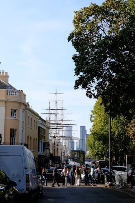 A busy street scene near Cutty Sark in Greenwich during daytime, featuring a large wooden sailing ship with multiple masts moored alongside modern buildings in the background. On the pavement, a group of pedestrians wait to cross, some carrying bags, while others are walking or standing. Close to the camera, there is a white van parked on the left side, likely used for home relocation or furniture transport by Man and a Van Greenwich, a professional removals company. Trees with green foliage line the street, casting partial shade on the scene, and a loading area is visible where furniture and boxes wrapped in plastic or cardboard are being moved, possibly being loaded onto or unloaded from the van. The scene captures the typical logistics involved in packing and moving, with visible equipment such as trolleys or straps, indicating an active process of house removals or relocation services in an urban environment. The lighting is natural, with bright daylight illuminating the scene, emphasizing the careful handling of belongings during a furniture transport or moving procedure.