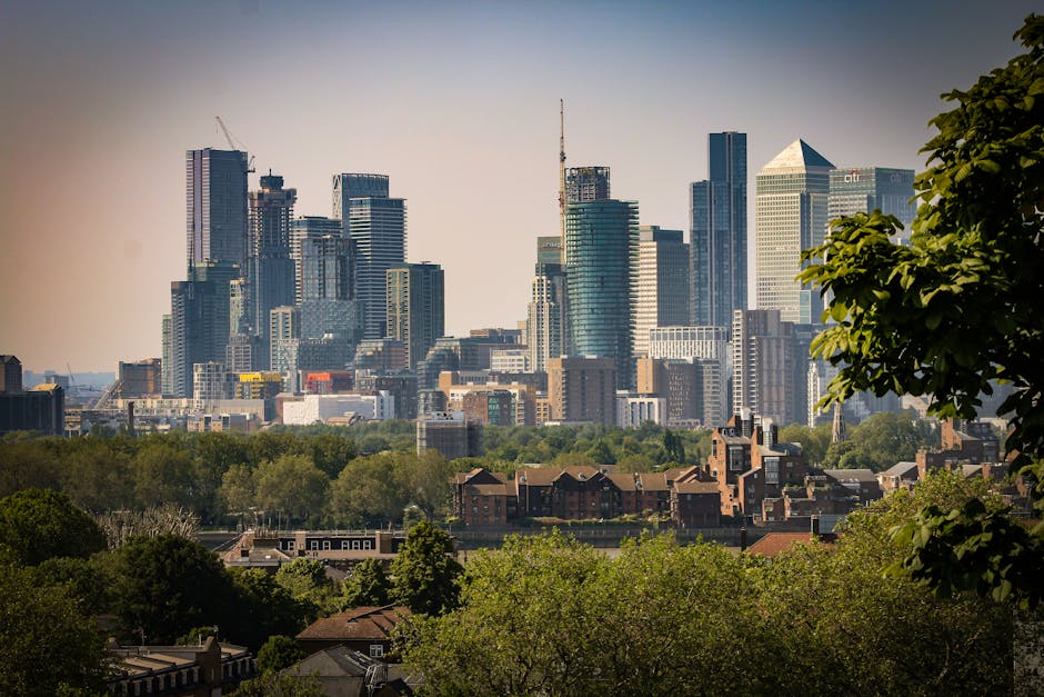 A city skyline featuring numerous modern skyscrapers, including glass high-rise buildings with reflective surfaces and various architectural designs, positioned behind a green area with dense trees an