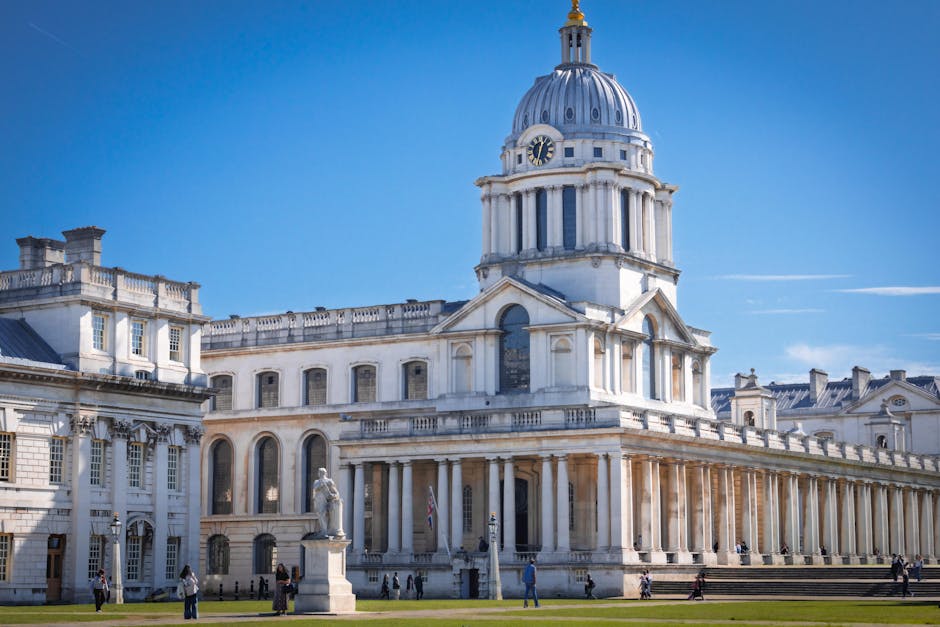 The image shows a historic white stone building with classical architectural features, including a large central dome topped with a golden ornament, columns, and arched windows. Prominent statues are positioned along the building's facade, and the structure is set against a clear blue sky. The scene includes people walking on the grass and steps in front of the building, with some individuals engaged in carrying or loading furniture and boxes, indicating a house removal or furniture transport process. Equipment such as trolleys and moving blankets are visible near the entrance, and a van is partially seen on the pavement, ready for loading or unloading. The environment appears to be part of a relocation or home moving operation, with the company's focus on packing and moving logistics, as seen in the preparation of household items for transportation. This setting reflects the professional loading process often involved in home relocation services provided by Man and a Van Greenwich, as they facilitate same-day removals in Greenwich and surrounding areas.