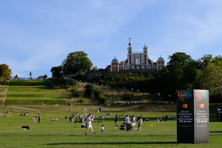 A view of a large, grassy park with multiple groups of people engaging in leisure activities such as sitting, walking, and playing with children and pets during daytime. In the background, a historic, multi-story building with ornate architectural details, domes, and a weather vane on top is situated atop a hill surrounded by trees. The park features tiered lawns and pathways, and there is a black freestanding sign advertising the Big Half marathon and offering a discount, positioned near the foreground on the right side of the image. The scene is well-lit with natural daylight, and the sky is partly cloudy with blue visible. This image, associated with house removals and moving services, captures the outdoor environment where relocation logistics like loading and unloading vehicles could take place.