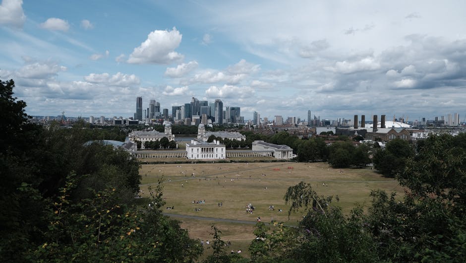 A wide view of Greenwich Park in London, with the National Maritime Museum and Queen's House in the foreground, surrounded by trees and open grassy areas with people relaxing and picnicking. Beyond the park, the London skyline features modern skyscrapers and high-rise buildings under a partly cloudy sky, creating a contrast between historic and contemporary architecture. The scene is captured from an elevated vantage point, possibly from a park pathway or vantage platform, during daytime with natural daylight illuminating the landscape. This setting illustrates the outdoor environment where a home relocation or furniture transport company like Man and a Van Greenwich might operate when assisting with house removals in the SE10 area, highlighting the transition from green spaces to urban logistics.